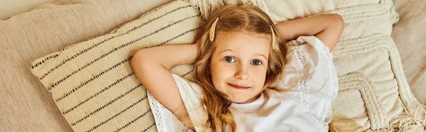 Chica alegre acostada en almohadas suaves con las manos detrás de la cabeza y mirando a la cámara en el dormitorio, pancarta - foto de stock
