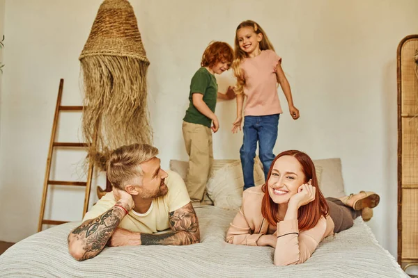 Joyeux mari et femme souriant près des enfants excités s'amuser sur le lit à la maison, temps de détente — Photo de stock