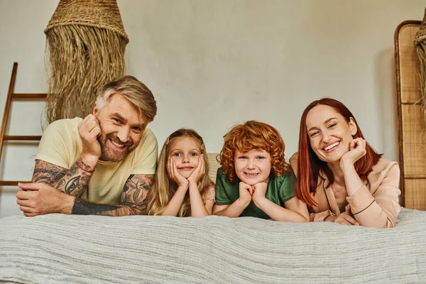 Freudige Eltern und unbeschwerte Kinder, die zu Hause auf dem Bett liegen und in die Kamera schauen, Erholungszeit — Stockfoto
