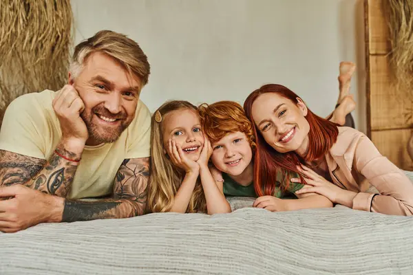 Parents heureux et enfants insouciants couchés sur le lit et regardant la caméra à la maison, temps de détente — Photo de stock