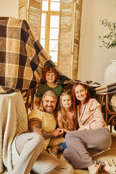 Padres y niños emocionados sentados debajo de una choza de mantas en la sala de estar, jugando juntos en casa - foto de stock