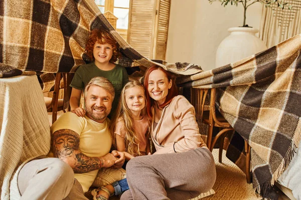 Padres alegres y niños riendo bajo la cabaña manta en la sala de estar, jugando juntos en casa - foto de stock