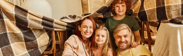 Cheerful couple with happy kids looking at camera under blanket hut in living room, banner — Stock Photo