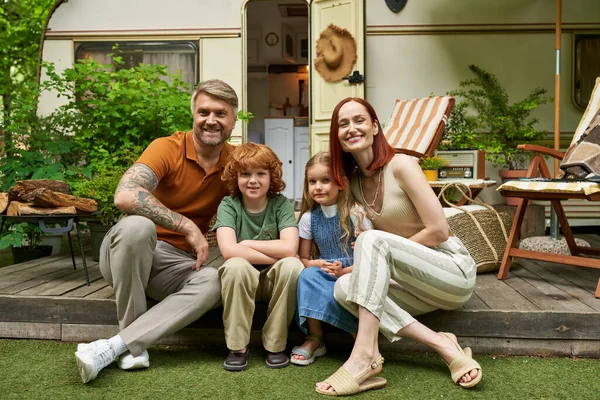 Fröhliche Eltern mit glücklichen Kindern, die in der Nähe ihres Wohnwagens sitzen und in die Kamera schauen, Familienfreizeit — Stockfoto