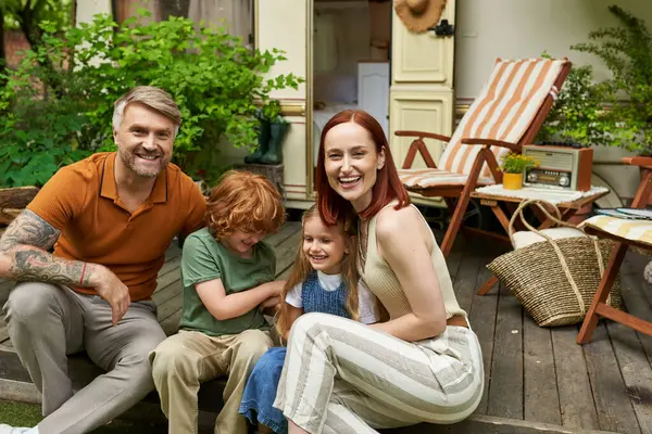 Padres riendo con niños felices sentados cerca de casa remolque y mirando a la cámara, tiempo en familia - foto de stock