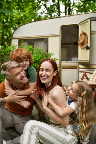 Niños alegres abrazando padres riendo sentados en casa remolque al aire libre, momentos de unión - foto de stock