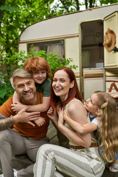 Crianças animadas abraçando pais rindo sentados em casa de reboque ao ar livre, conexão emocional — Fotografia de Stock