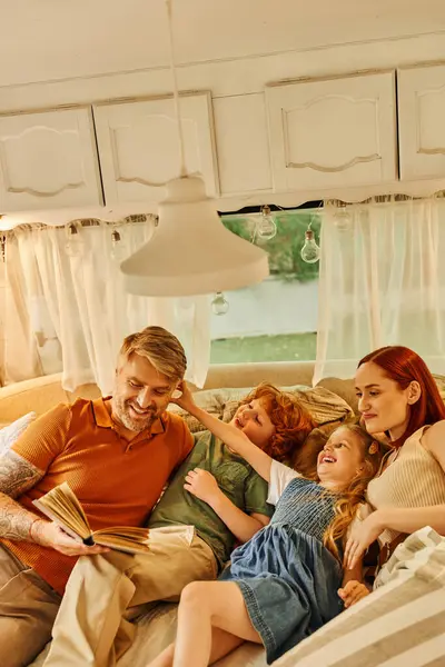 Fröhlich tätowierter Mann liest Familie Buch auf weichem Bett im gemütlichen Wohnwagen vor, lernt gemeinsam — Stockfoto