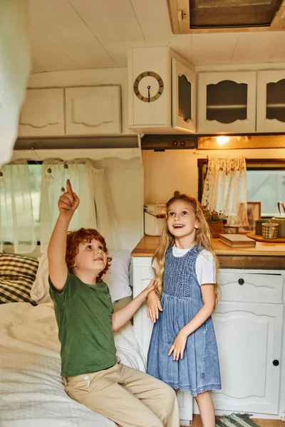 Pelirroja apuntando hacia arriba en casa remolque con interior acogedor moderno cerca de hermana sorprendida, hermanos felices - foto de stock