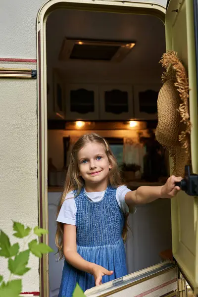 Rubia chica en denim vestido ventana de apertura en casa remolque y mirando a la cámara, feliz infancia - foto de stock