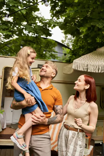 Happy tattooed man holding cute daughter near smiling wife and trailer home on park, leisure — Stock Photo