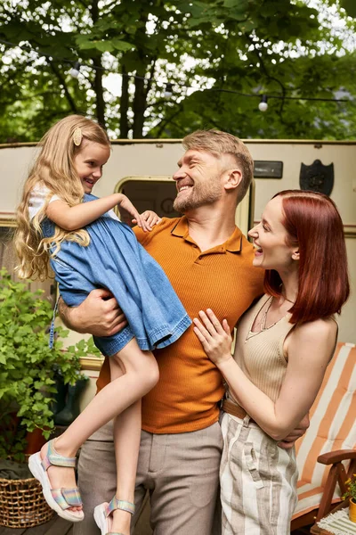 Alegre homem segurando bonito filha perto sorrindo esposa e reboque casa no parque, ligação familiar — Fotografia de Stock