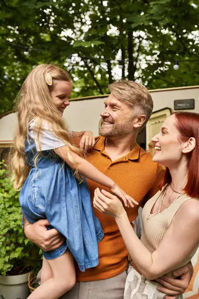Father holding cheerful daughter near pleased wife and modern trailer home, leisure and adventure — Stock Photo