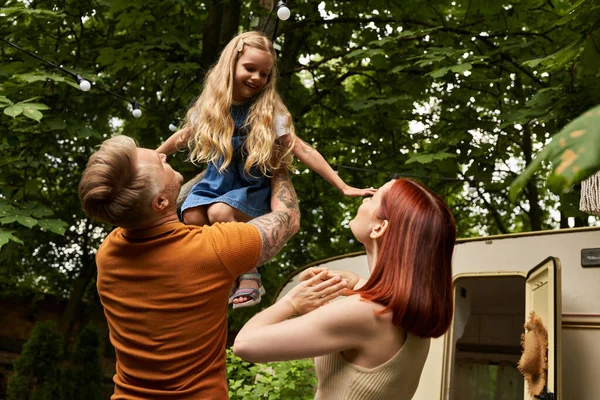 Tatuato uomo holding overjoyed figlia in mani vicino moglie e moderno rimorchio casa, famiglia tempo libero — Foto stock