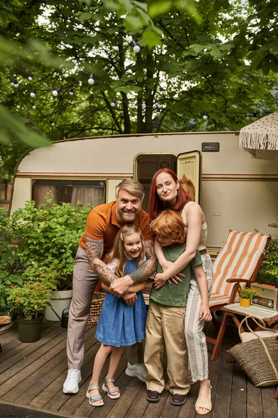 Happy parents embracing cheerful children near modern home on wheels in trailer park, family leisure — Stock Photo