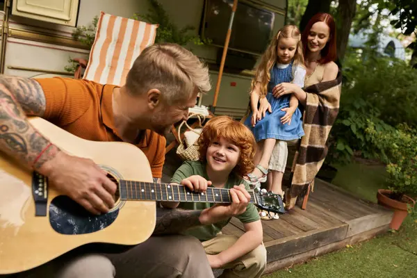 Hombre tatuado tocando la guitarra acústica alegre pelirroja niño cerca de la familia y remolque moderno a casa - foto de stock