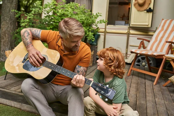 Hombre tatuado tocando la guitarra acústica a hijo pelirrojo junto a casa remolque al aire libre, diversión y aprendizaje - foto de stock