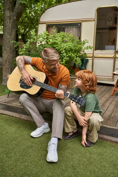 Padre tatuato che suona la chitarra acustica al figlio sorridente accanto alla casa del rimorchio, divertimento e apprendimento — Foto stock
