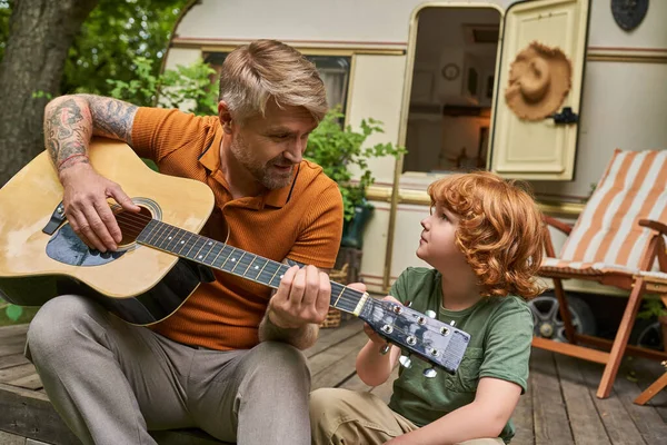 Tätowierter Vater spielt dem aufmerksamen Sohn neben Wohnwagen Akustikgitarre, Spaß und Lernen — Stockfoto