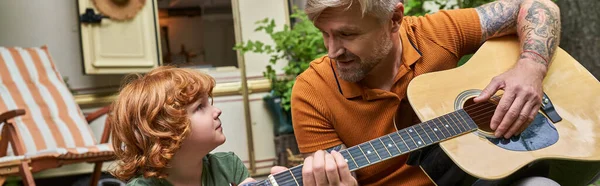 Hombre feliz tocando la guitarra acústica al hijo atento al lado del remolque a casa, diversión y aprendizaje, bandera - foto de stock