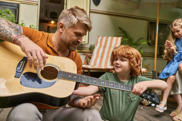 Tatuado hombre enseñando pelirroja hijo a tocar la guitarra acústica cerca de casa sobre ruedas en remolque parque - foto de stock