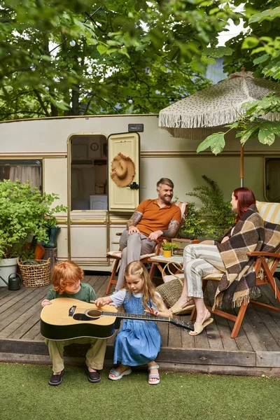 Kinder lernen Akustikgitarre spielen, während Eltern in Liegestühlen in der Nähe ihres Wohnwagens sitzen — Stockfoto