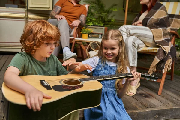 Pelirrojo chico aprender a tocar la guitarra cerca alegre hermana y padres sentado en casa remolque - foto de stock