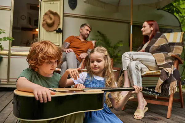 Redhead child learning to play guitar near sister and parents sitting and talking at trailer home — Stock Photo