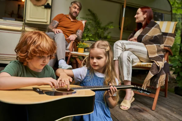 Lindos hermanos aprendiendo a tocar la guitarra cerca de los padres sentados en hablar al lado de casa remolque - foto de stock