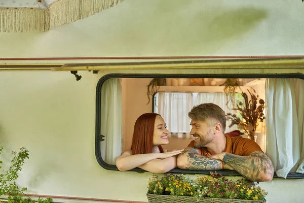 Heureux tatoué homme et rousse femme regarder l'autre dans fenêtre de moderne remorque maison — Photo de stock