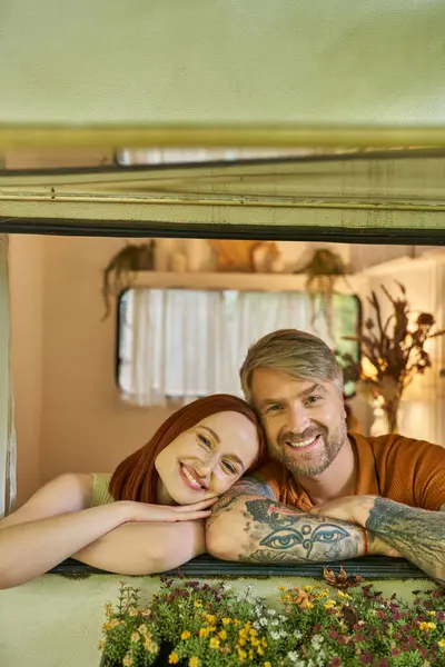 Cheerful redhead woman and tattooed man looking at camera from window of modern trailer home — Stock Photo
