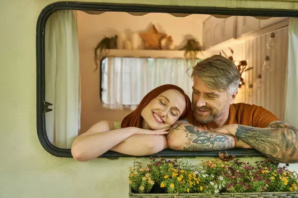 Feliz mulher ruiva sorrindo com os olhos fechados perto de homem tatuado na janela da moderna casa reboque — Fotografia de Stock