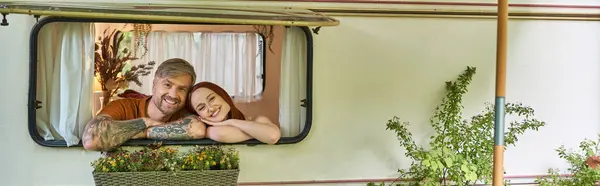Возбужденная и стильная пара, смотрящая в окно мобильного дома вокруг зелени в трейлерном парке, баннер — стоковое фото