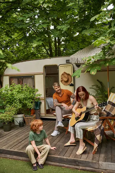 Mulher tocando guitarra acústica perto de marido tatuado e filho ruivo sentado em casa trailer moderno — Fotografia de Stock