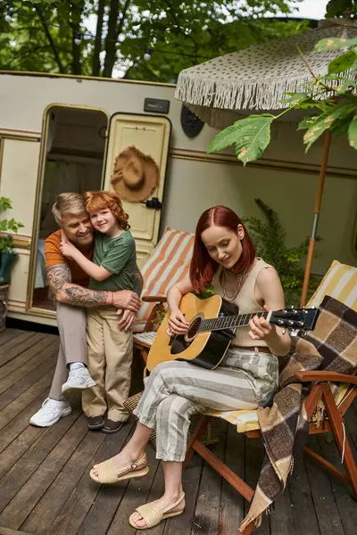 Gioiosa donna suonare la chitarra acustica vicino tatuato marito abbracciare figlio accanto al rimorchio casa — Foto stock