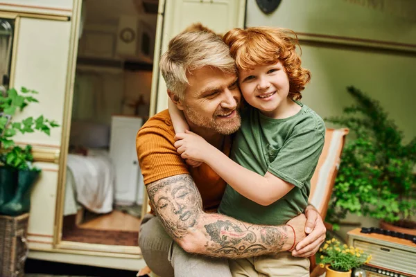 Feliz tatuado homem abraçando adorável ruiva filho perto moderna reboque casa, momentos de ligação — Fotografia de Stock