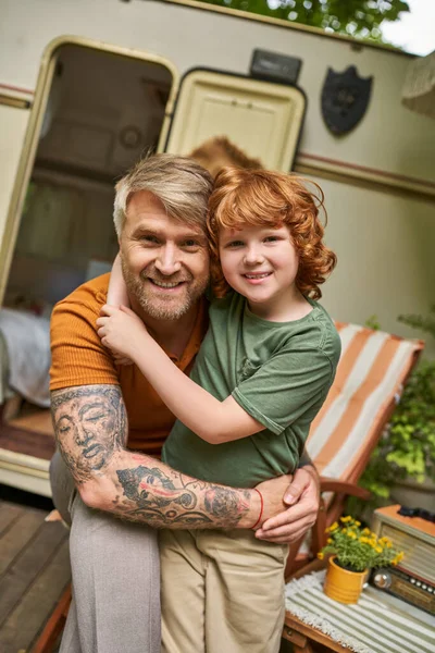 Freudig tätowierter Mann mit entzückendem rothaarigem Sohn, der sich neben Wohnwagen umarmt und in die Kamera lächelt — Stockfoto