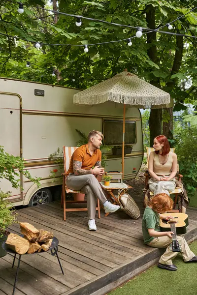 Redhead boy playing acoustic guitar while parents sitting and talking next to moder trailer home — Stock Photo