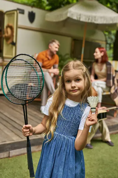 Girl showing badminton rockets and shuttlecock near family and trailer home on blurred background — Stock Photo
