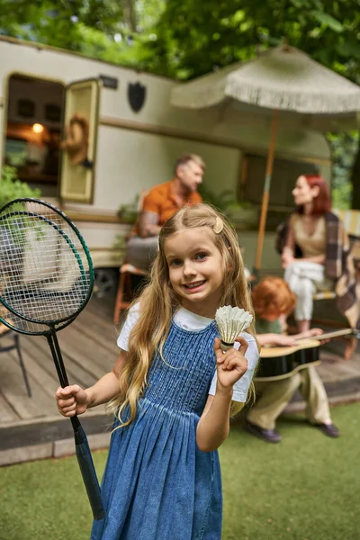 Menina feliz com badminton foguetes e shuttlecock perto da família e reboque casa no fundo borrado — Fotografia de Stock