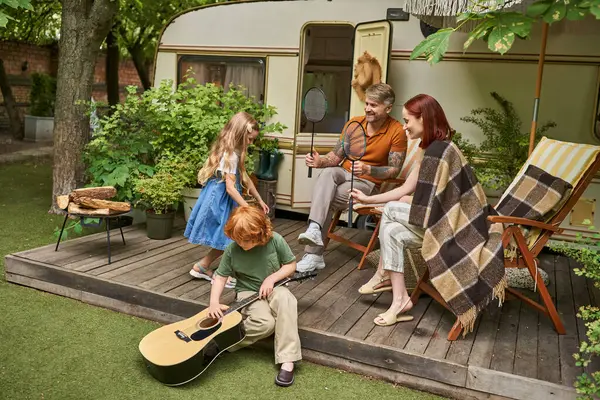 Boy learning to play guitar near sister and parents with badminton rockets next to trailer home — Stock Photo
