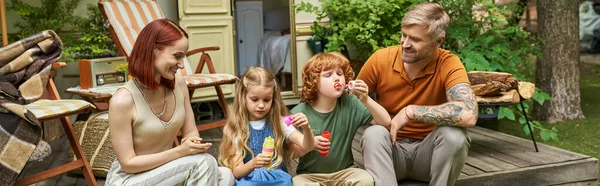 Smiling parents looking at children blowing soap bubbles near trailer home, leisure and fun, banner — Stock Photo