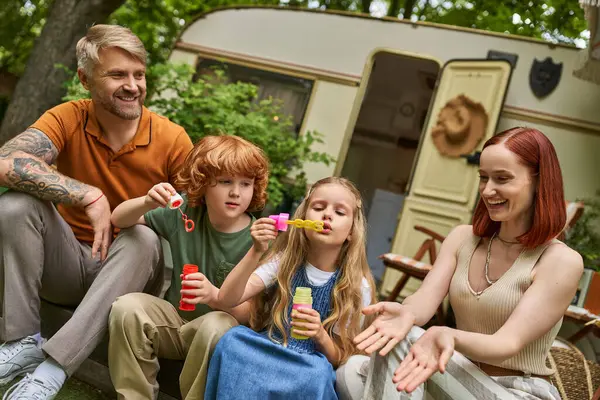 Simpatici bambini che soffiano bolle di sapone vicino ai genitori felici accanto alla moderna casa rimorchio, tempo libero e divertimento — Foto stock
