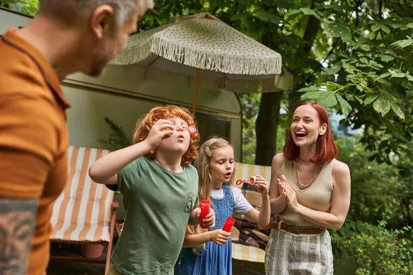 Niños soplando burbujas de jabón cerca de los padres riendo al lado de casa remolque moderno, ocio y diversión - foto de stock