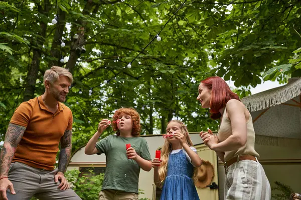 Smiling parents looking at adorable children blowing soap bubbles near trailer home outdoors — Stock Photo