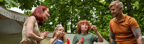 Pais alegres olhando para crianças bonitos soprando bolhas de sabão perto de casa reboque ao ar livre, banner — Fotografia de Stock