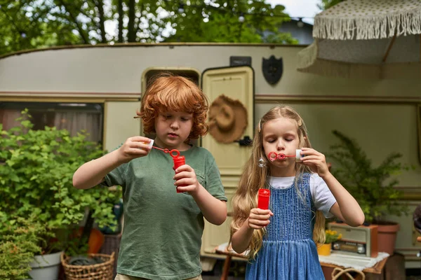 Pelirrojo chico y rubia chica soplando burbujas de jabón cerca de casa remolque, hermanos jugando juntos - foto de stock