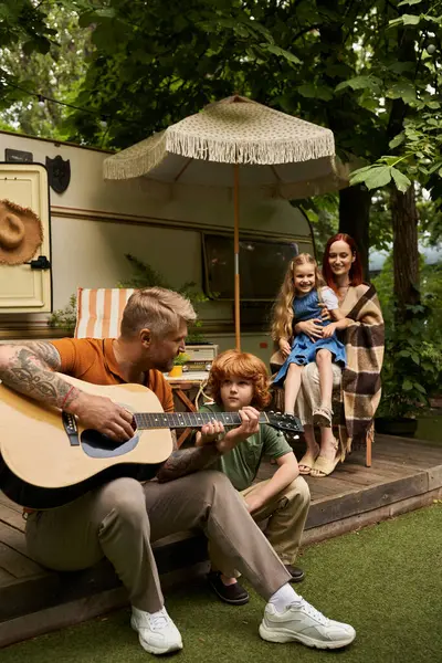 Hombre tatuado tocando la guitarra al hijo pelirrojo atento y la familia sonriendo junto al remolque a casa - foto de stock