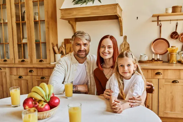 Счастливые родители с милой дочерью, глядя в камеру рядом со свежими фруктами и апельсиновым соком на кухне — стоковое фото