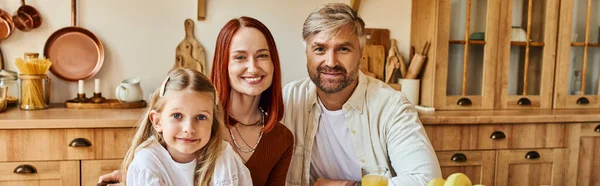 Pais sorridentes com filha adorável sorrindo para a câmera na cozinha acolhedora em casa, bandeira horizontal — Fotografia de Stock